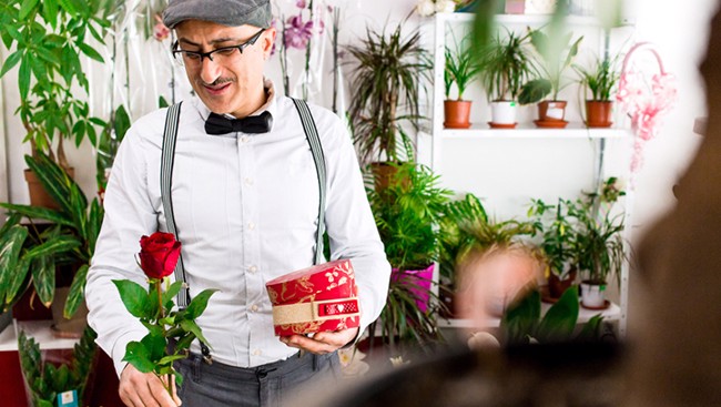 Een man in een bloemenwinkel houdt een rode roos en een ronde cadeauverpakking vast, omringd door groene planten.