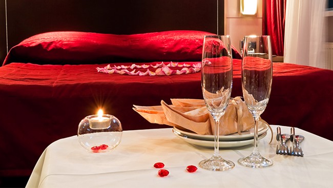 Een romantisch gedekte tafel met twee champagneglazen, een brandend kaarsje en broodmandje staat voor een bed met rood beddengoed en rozenblaadjes.