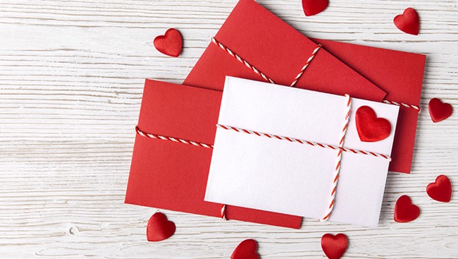 Drie enveloppen in rood en wit met touw en hartversieringen liggen op een witte houten ondergrond, met kleine rode hartjes eromheen.