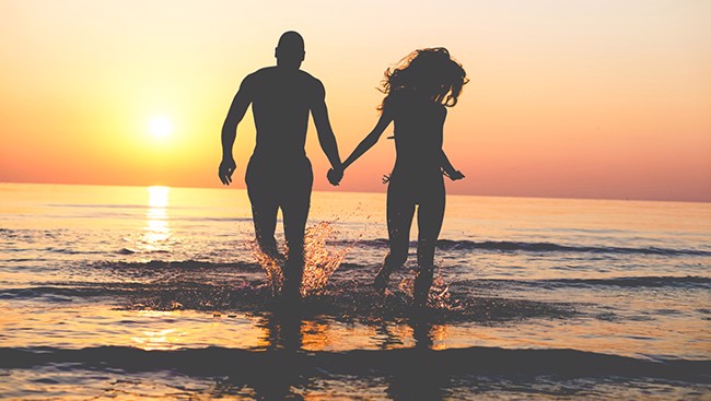 Een stel loopt hand in hand door het ondiepe water bij zonsondergang, met de zon laag aan de horizon en warme oranje gloed over zee.