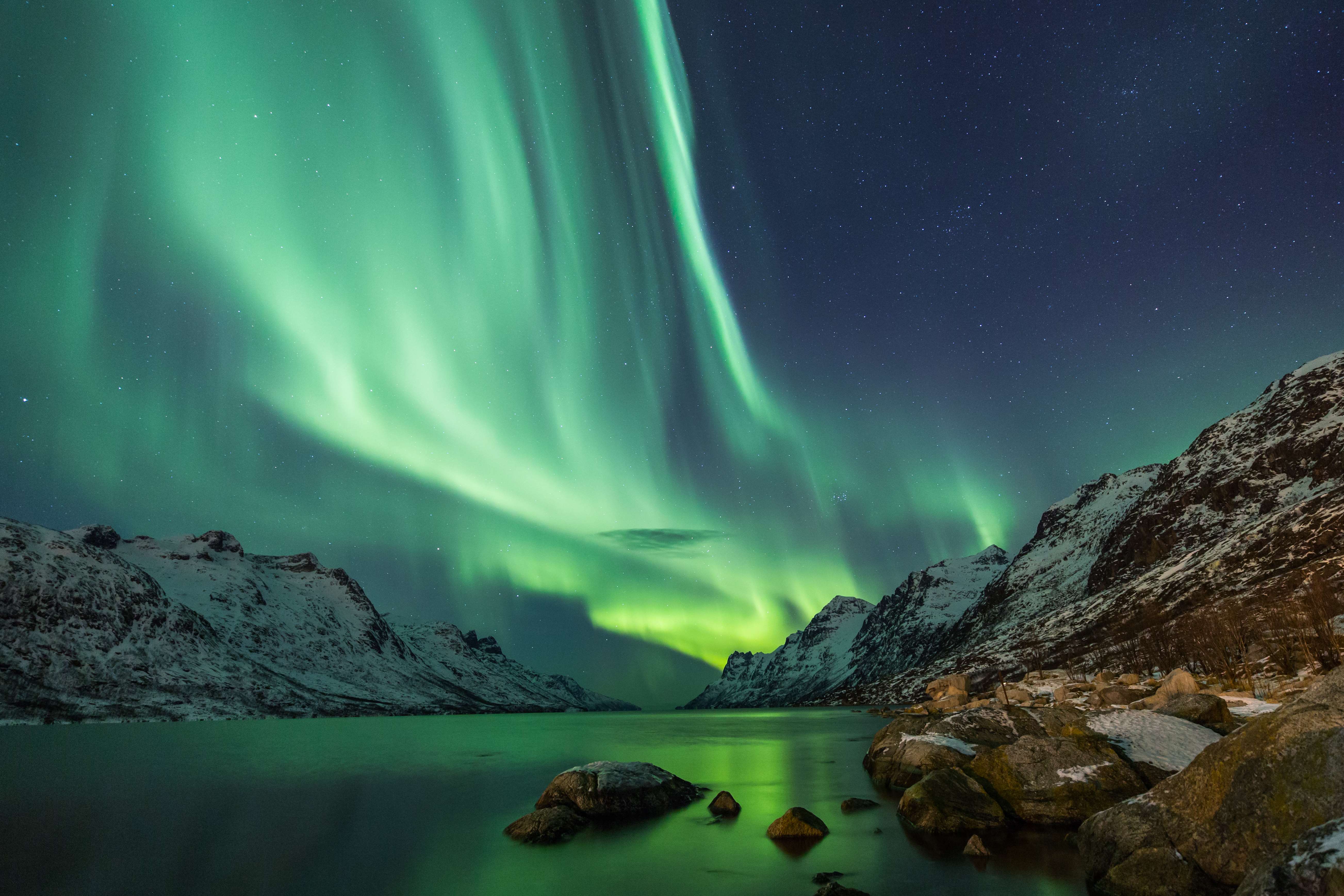 Groen noorderlicht danst boven een besneeuwde fjord in Finland, met donkere bergen, rotsige oevers en een stille reflectie in het water onder de nachtelijke hemel.