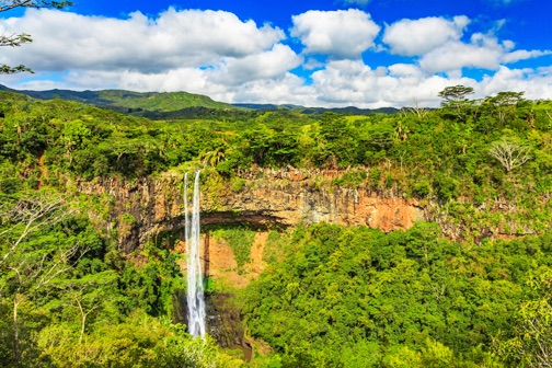 Hoge waterval die langs een groene klif naar beneden stroomt, omringd door dicht tropisch regenwoud en heuvels van Mauritius onder een blauwe lucht.