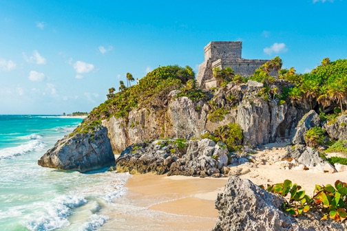 Rotsachtige baai met zandstrand en turquoise zee, met groene begroeiing en oude tempelruïnes bovenop de klif aan de Mexicaanse kust.