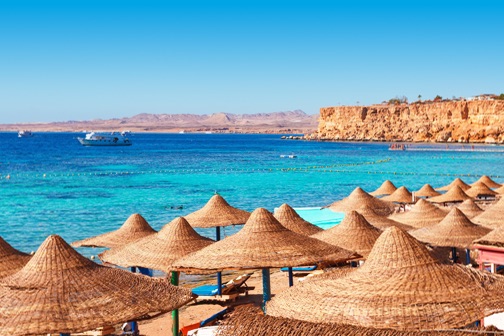 Rieten parasols op een zonnig strand aan helderblauw water, met rotsachtige kust en zee op de achtergrond in Egypte.