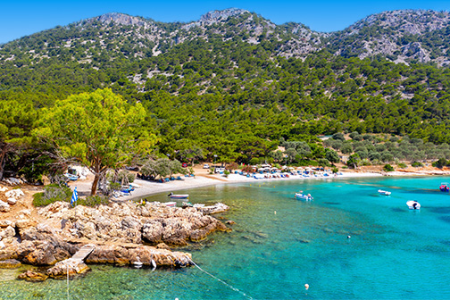 Baai op Samos met helder turquoise water, rotsachtige kust en klein strand met ligbedden, omringd door groene heuvels en bergen.