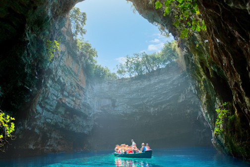 Ronde grot met open dak waar zonlicht binnenvalt, helderblauw water en een bootje met mensen in het midden van de lagune.