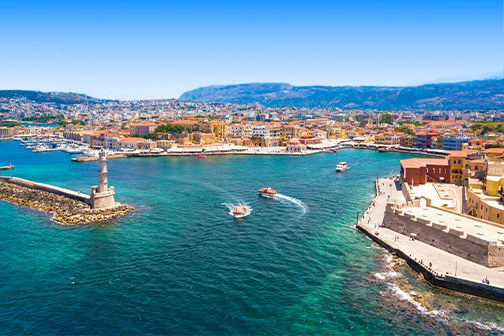 Havenstad Chania met vuurtoren op de pier, turquoise water met bootjes en kleurrijke gebouwen langs de kade onder een heldere lucht.