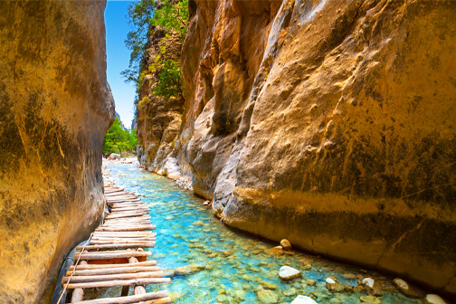 Smalle kloof met hoge rotswanden en helder turquoise water, met een houten loopbrug langs de rand van de rivier.