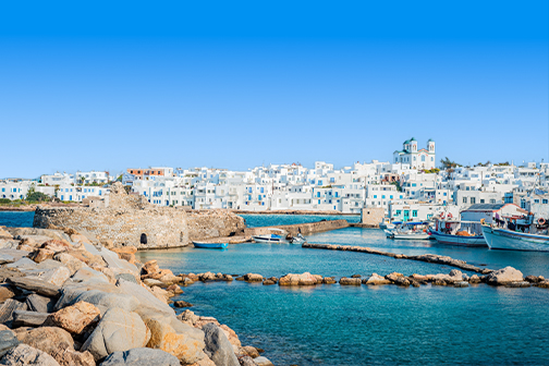 Haven van Naoussa met turquoise water en vissersboten, voor een stad met witte huizen en een kerk met blauwe koepel onder heldere lucht.