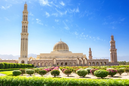 Grote moskee met centrale koepel en hoge minaretten, omringd door verzorgde tuinen en bloemen onder een heldere blauwe lucht in Muscat.
