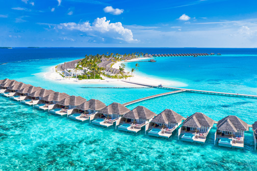 Watervilla’s op palen boven helder turquoise water, verbonden door een houten pier naar een groen eiland, gezien vanuit de lucht op de Malediven.