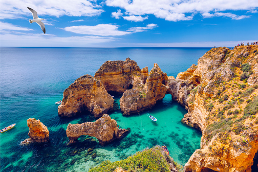 Rotsachtige kust met hoge kliffen en natuurlijke bogen boven helder turquoise water; kleine bootjes en een meeuw onder blauwe lucht.