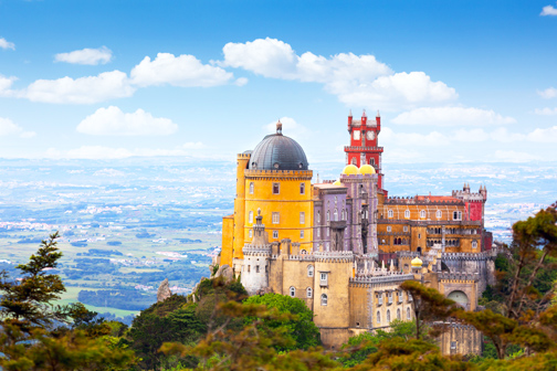 Kleurrijk paleis in Portugal op een beboste heuvel, met gele gevels, koepel en torens, uitkijkend over een groen landschap onder een blauwe lucht.