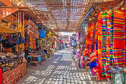 Smalle steeg in een kleurrijke souk met kramen vol stoffen, tassen en souvenirs, waar zonlicht door een rieten dak naar binnen valt in Marrakech.