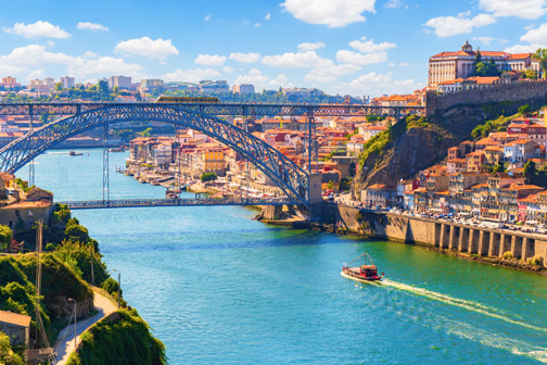 Breed uitzicht op een rivier in Porto met een grote stalen boogbrug, kleurrijke huizen langs de oevers en een boot die door het blauwe water vaart.