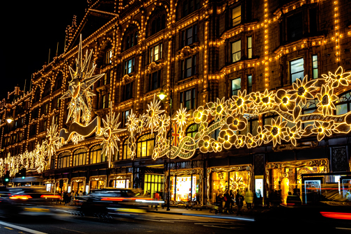 Groot warenhuis in Londen vol warme feestverlichting aan een drukke straat, met lichtgevende sterren en bloemen terwijl voorbijrijdende auto’s strepen van beweging vormen.