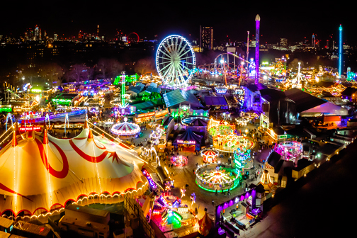 Fel verlicht kermisterrein in Londen bij nacht met een groot reuzenrad, kleurrijke attracties en tenten, omringd door bezoekers in een levendige stadse setting