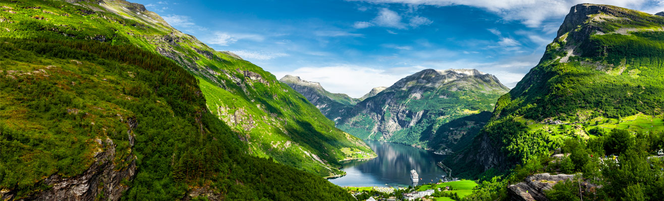 Groene fjordenvallei in Noorwegen met steile bergen en een glinsterend meer, waar een cruiseschip en huizen langs de oever liggen onder een helderblauwe lucht.