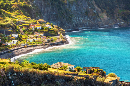 Kustlijn van Madeira met steile rotsen, een dorpje langs de baai en helderblauwe zee, gezien vanaf een hoger gelegen punt.