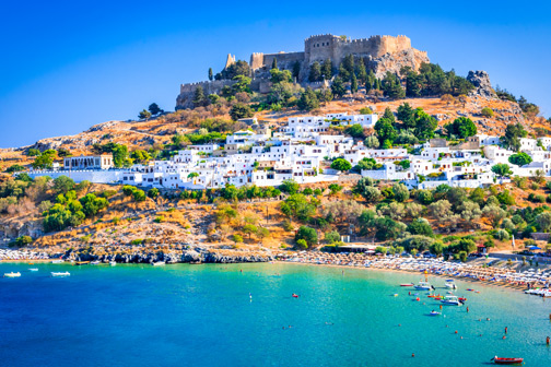 Witte huizen tegen een heuvel aan zee, met bovenop een oud akropolis en een baai met helderblauw water en kleine bootjes.