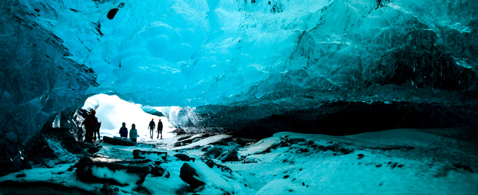 Groepswandelaars lopen door een blauw verlichte ijsgrot in IJsland, met dikke ijswanden, sneeuw op de grond en licht dat via de opening naar binnen valt.