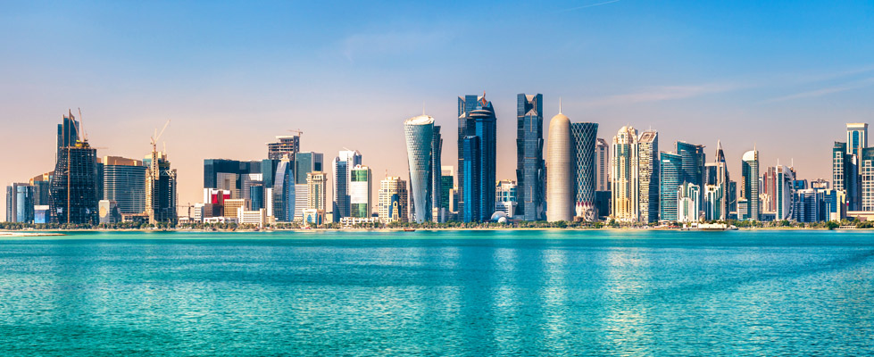 Moderne skyline van Qatar met hoge glazen torens langs een rustige turquoise baai onder een zachte, helderblauwe lucht.