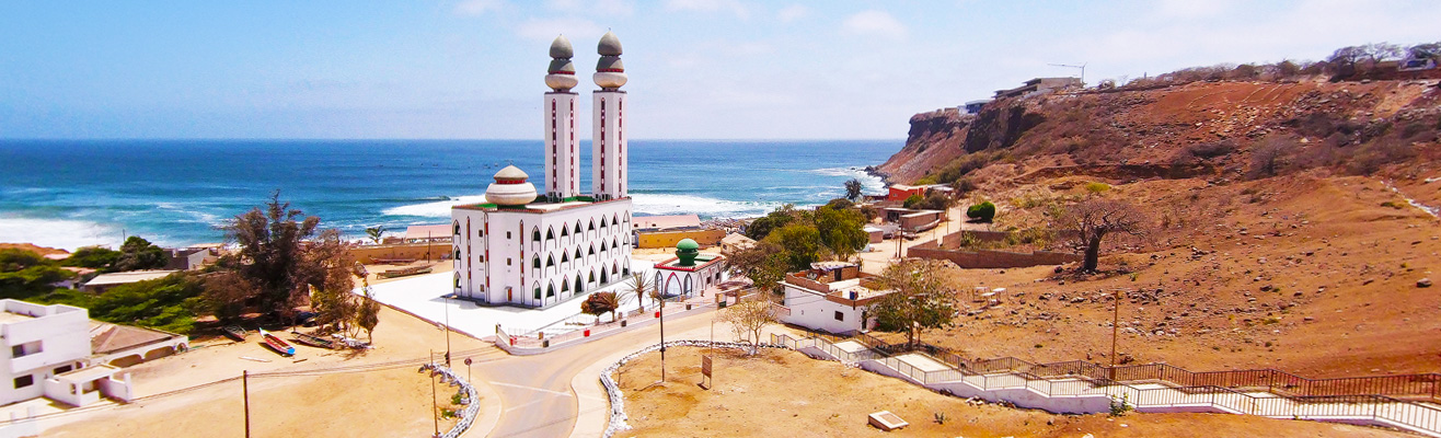 Witte kustmoskee met hoge minaretten aan de rand van een rotsachtig landschap in Senegal, uitkijkend over de blauwe oceaan en golvende kustlijn.