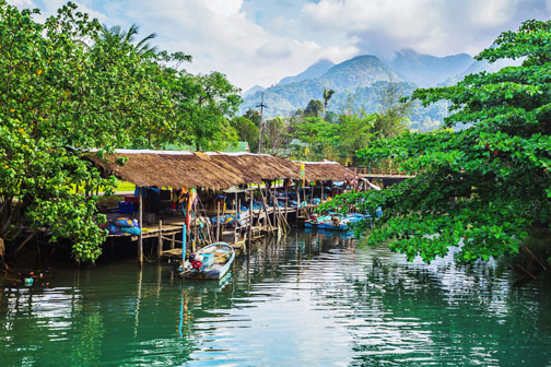 Tropisch rivierlandschap in Thailand met houten huisjes op palen, vissersboten en groene bergen op de achtergrond.