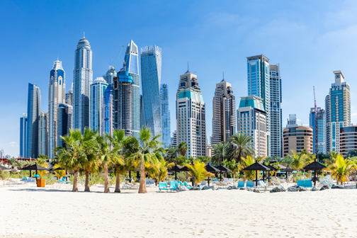 Zandstrand in Dubai met palmbomen en ligbedden, met op de achtergrond een moderne skyline van hoge wolkenkrabbers onder een heldere blauwe lucht.