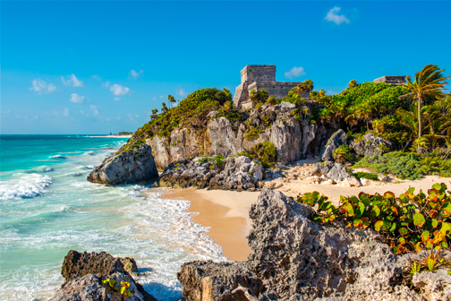 Rotsachtige baai in Mexico met zandstrand en turquoise golven, palmen en groene begroeiing, met oude tempelruïnes bovenop de klif.