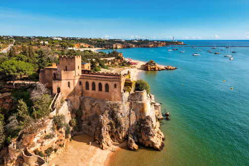 Kasteel op een rotsachtige klif aan zee in de Algarve, met zandstrand eronder en helderblauw water met zeilboten langs de kust.