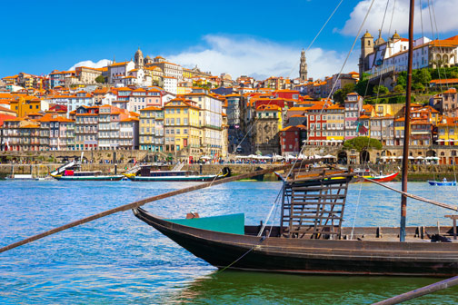 Traditionele houten boot op een rivier, met op de achtergrond een heuvel vol kleurrijke huizen en historische gebouwen onder een heldere blauwe lucht.