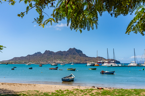 Rustig strand op Kaapverdië aan een helderblauwe baai met kleine bootjes voor anker en zeiljachten verderop, met groene heuvels op de achtergrond.