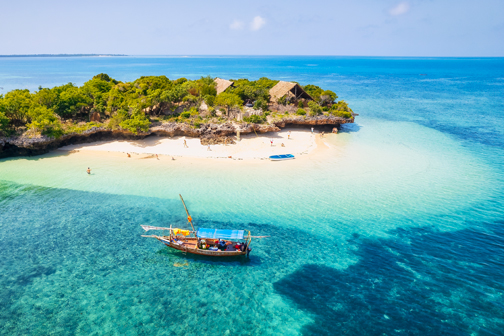 Helderblauwe zee rond een klein eiland met wit zandstrand en groene begroeiing, met een houten boot op het rustige, ondiepe water bij Zanzibar.