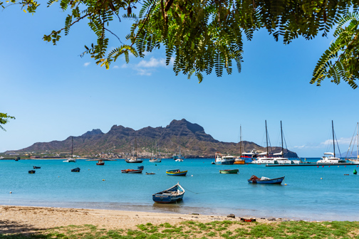 Rustig strand op Kaapverdië aan een helderblauwe baai met kleine bootjes voor anker en zeiljachten verderop, met groene heuvels op de achtergrond.